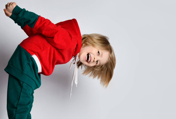 Portrait of happy smiling playful frolic blond kid boy in dark gray and red hoodie and pants bending over - Photo, Image
