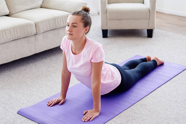 Portrait of young woman practicing yoga at home indoor, copy space. Girl practicing cobra pose, full length. Relaxing and doing yoga. Wellness and healthy lifestyle. Bhujangasana - Photo, image