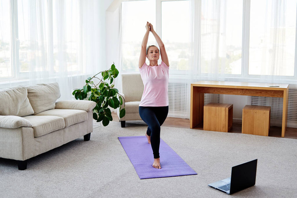 Portrait of young yogini woman practicing balance yoga asana Virabhadrasana at home, copy space. Online training on laptop computer. Yoga, pilates, working out exercising - Foto, Bild