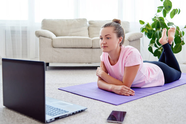 Beautiful young woman doing exercise on floor at home, online training on laptop computer, copy space. Full length portrait. Yoga, pilates, working out exercising - Foto, Bild