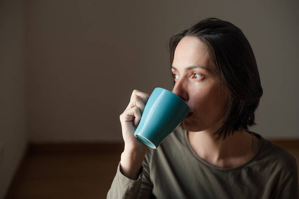 穏やかな幸せな表情で居心地の良い朝に自宅でコーヒーを飲む若い自然美女性. - 写真・画像