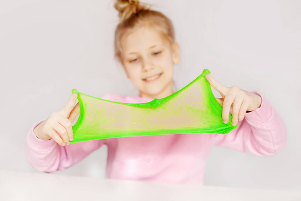 Χαριτωμένο κορίτσι που παίζει με πράσινη γλίτσα. Το κορίτσι κρατά στα χέρια της και τεντώνει τη βλέννα στη διαφάνεια. Slime παιχνίδι, αντι-στρες παιχνίδι - Φωτογραφία, εικόνα