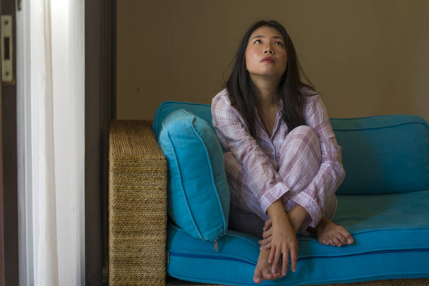 indoors lifestyle portrait of young beautiful sad and depressed Asian Korean woman in pajamas at home sitting by living room couch feeling unhappy and confused suffering broken heart pain - Fotoğraf, Görsel