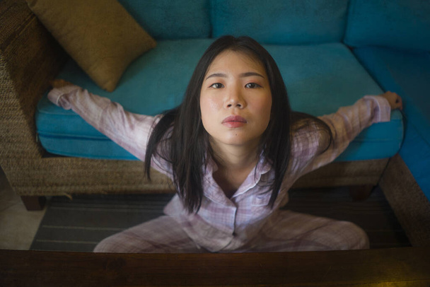 dramatic dark portrait of young beautiful sad and depressed Asian Chinese woman in pajamas at home sitting by living room couch feeling unhappy and confused suffering broken heart pain - Zdjęcie, obraz