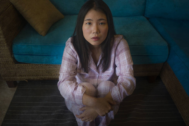 Evinde pijamalarıyla oturan üzgün ve üzgün Asyalı genç bir kadının dramatik karanlık portresi. Oturma odasındaki kanepede oturuyor. Mutsuz ve kafası karışık, kırık kalp ağrısı çekiyor. - Fotoğraf, Görsel