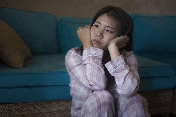dramatic dark portrait of young beautiful sad and depressed Asian Chinese woman in pajamas at home sitting by living room couch feeling unhappy and confused suffering broken heart pain - Foto, afbeelding