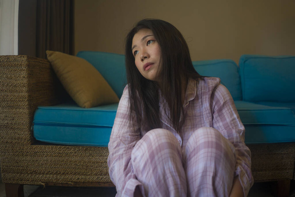 indoors lifestyle portrait of young beautiful sad and depressed Asian Japanese woman in pajamas at home sitting by living room couch feeling unhappy and confused suffering broken heart pain - Foto, Bild