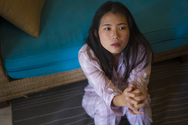 indoors lifestyle portrait of young beautiful sad and depressed Asian Japanese woman in pajamas at home sitting by living room couch feeling unhappy and confused suffering broken heart pain - Zdjęcie, obraz