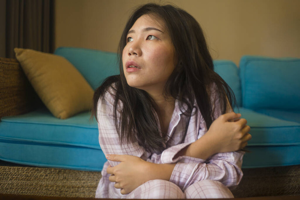 indoors lifestyle portrait of young beautiful sad and depressed Asian Korean woman in pajamas at home sitting by living room couch feeling unhappy and confused suffering broken heart pain - Фото, зображення