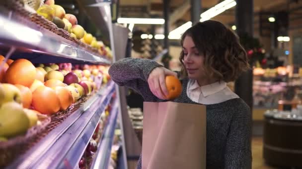 Cute girl buys fresh oranges in the market. Beautiful young woman stands in front the shelf and puts the oranges to a brown paper bag, she is pleased with the choice - Video