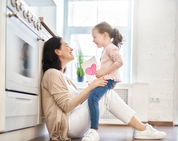 Happy mother's day! Child daughter is congratulating mom and giving her postcard. Mum and girl smiling and hugging. Family holiday and togetherness.  - 写真・画像