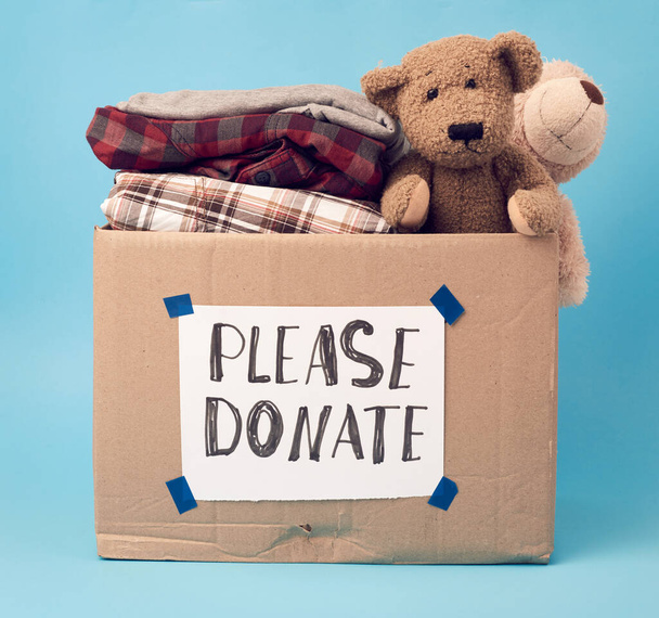 nagy barna kartondoboz felirattal kérjük adományozni, doboz tele van különböző ruhák, játékok a kék háttér - Fotó, kép