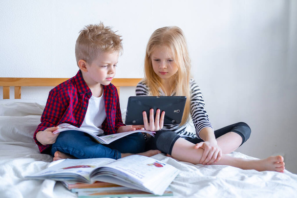 Дистанционное обучение онлайн. Школьник и девочка учатся дома с цифровым планшетным ноутбуком и выполняют домашнюю работу в школе. Сидя на кровати с учебниками
. - Фото, изображение