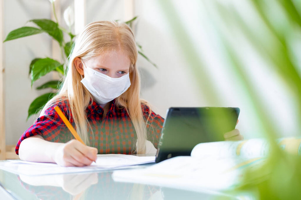 Distance learning online education. Sickness schoolgirl in medical mask studying at home with digital tablet in hand and doing school homework. Training books and notebooks on table. - Fotó, kép