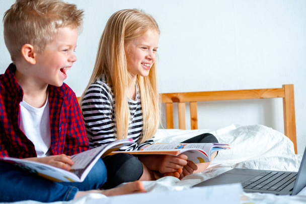 遠隔オンライン教育。学校の男の子と女の子は、デジタルタブレットノートブックで自宅で勉強し、学校の宿題を行う。トレーニング本と一緒にベッドに座って. - 写真・画像