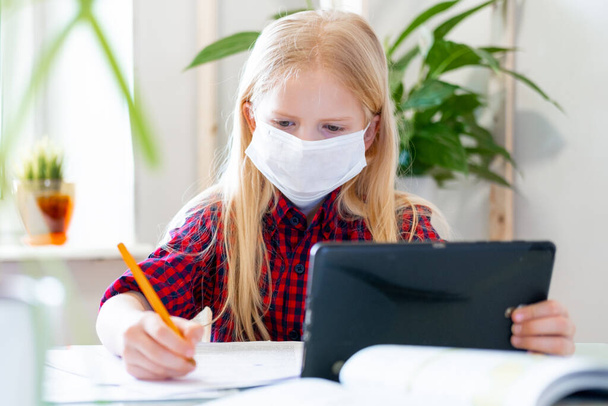 Distance learning online education. Sickness schoolgirl in medical mask studying at home with digital tablet in hand and doing school homework. Training books and notebooks on table. - Φωτογραφία, εικόνα