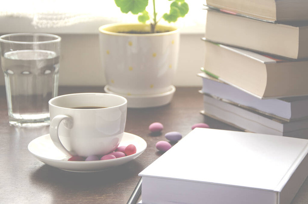 The white paper lies on a wooden table next to a stack of colorful books, a cup of coffee and a pot against the background of the window - Photo, Image