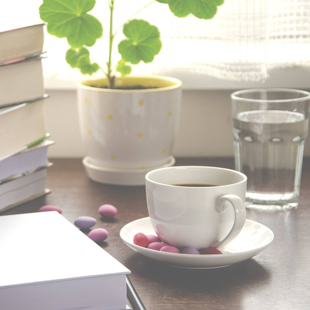 The white paper lies on a wooden table next to a stack of colorful books, a cup of coffee and a pot against the background of the window - Foto, immagini