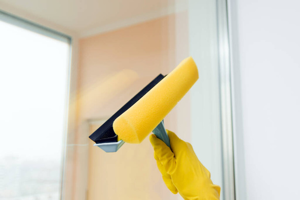 Window cleaner for washing a windows. Hand in yellow glove hold cleaning scraper. Cleaning a glass with a squeegee, close up. - 写真・画像