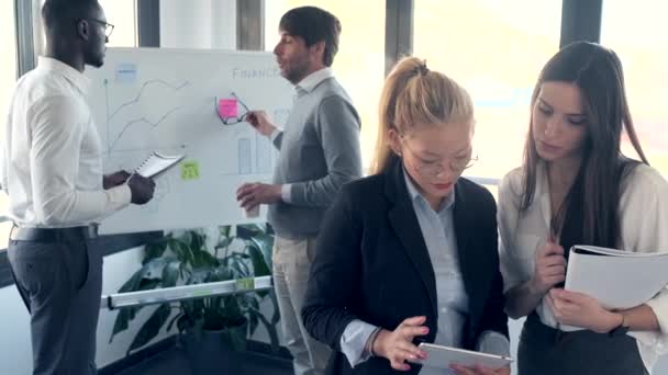 Twee elegante jonge zakenvrouw werken met digitale tablet terwijl haar collega 's uitleggen het project in de voorkant van whiteboard in coworking ruimte. - Video