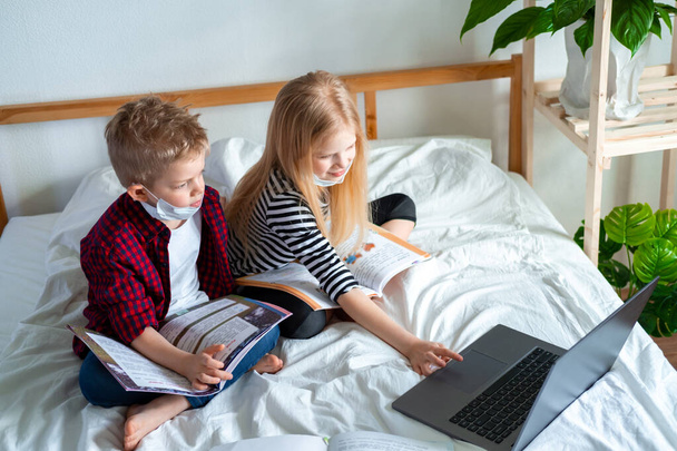 Distance learning online education. Sickness school boy and girl in medical mask studying at home with digital tablet laptop notebook and doing homework. Sitting on bed with training books - Fotó, kép