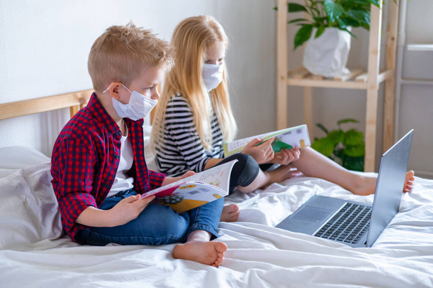 Distance learning online education. Sickness school boy and girl in medical mask studying at home with digital tablet laptop notebook and doing homework. Sitting on bed with training books - Photo, image