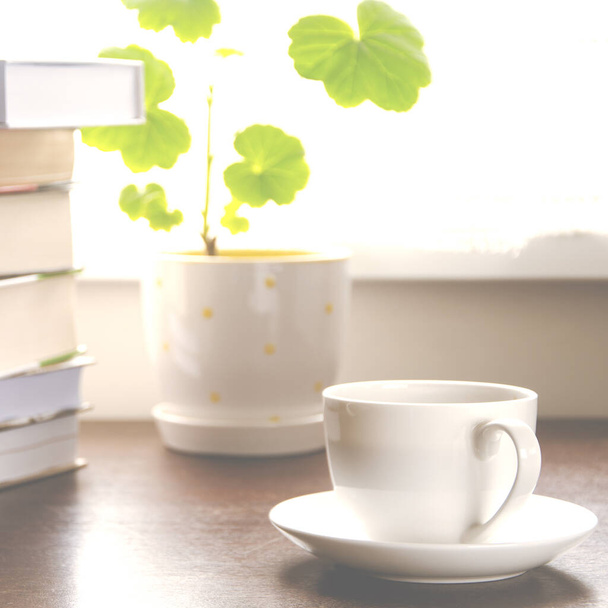 A white cup of coffee stands on a wooden table next to a stack of books and a pot against the background of the window. Close-up - Foto, Bild