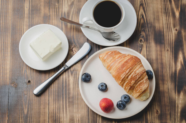 Coffee cup, croissant with berries in white bowl and butter knife on wooden table. Top view. Healthy breakfast with fresh berries - Фото, изображение