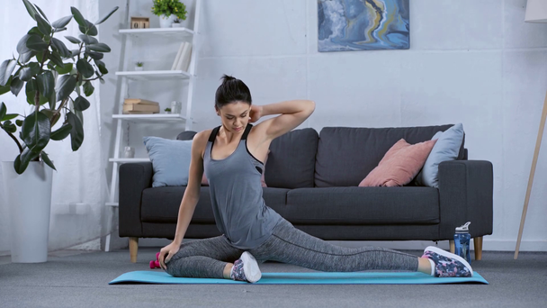 Side view of sportswoman stretching on fitness mat in living room - Filmati, video