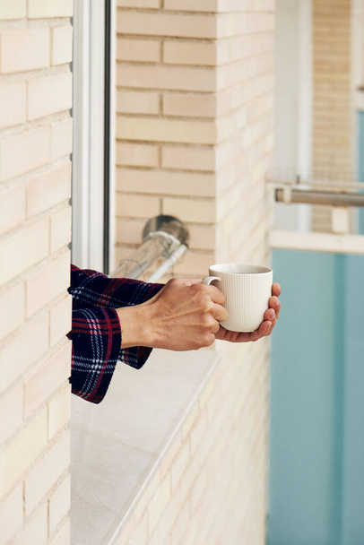 closeup of a young caucasian man, in pajamas, having a coffee leaning on the railing of a window, at his house or of a hotel - Photo, Image