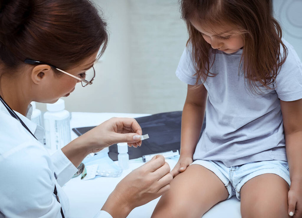 Cute little baby girl at the pediatrician's appointment, nice young woman doctor in a white medic lab coat with gentleness glues a band-aid on girl's knee, child in the hospital - Photo, image