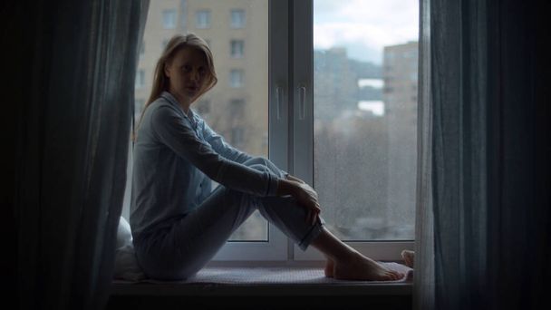 Nuori kaunis nainen kotivaatteissa ja suojaava naamio istuu ikkunalaudalla ja katselee ulos ikkunasta. Karanteeni sepelvaltimotautiepidemian aikana. COVID-19-pandemia
 - Valokuva, kuva
