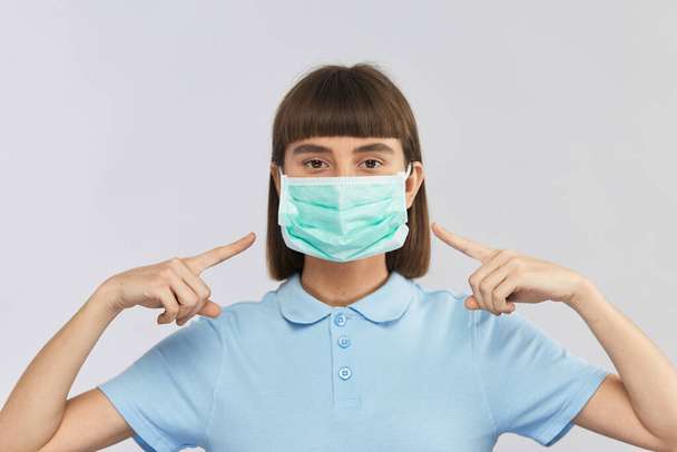 νεαρή σοβαρή κοπέλα φορώντας αποστειρωμένη μάσκα προστασίας και poiting με τα δάχτυλα σε αυτό, προστασία πρέπει να είναι ενώ coronavirus - Φωτογραφία, εικόνα