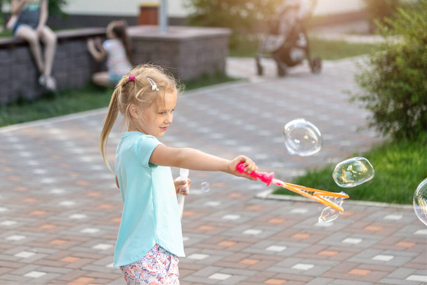 Porträt eines kleinen süßen kaukasischen blonden Mädchens, das Spaß und Freude beim Blasen großer Seifenblasen im Stadtpark hat. Fröhlich liebenswert Kind genießen Sie gesunde Aktivitäten außerhalb des Sommers Spiel. - Foto, Bild