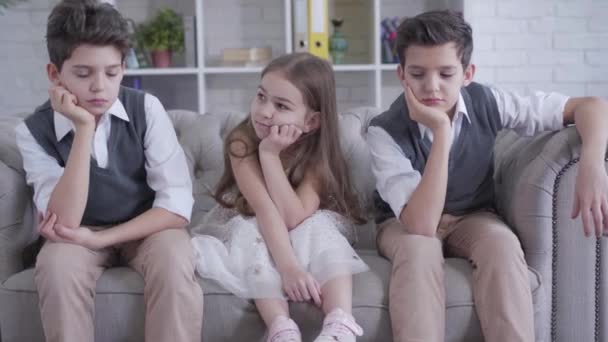 Caucasian twin brothers sitting on couch on both sides of cheerful cute girl. Portrait of bored children resting indoors. Friendship, childhood, leisure, lifestyle. - Video