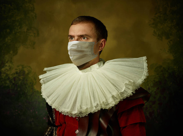 Young man as a medieval knight on dark background wearing protective mask against coronavirus - 写真・画像