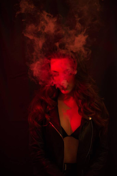 Young attractive girl with long curly hair smokes an electronic cigarette and releases smoke in the red spotlights. - 写真・画像