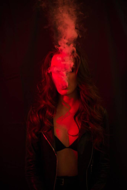 Young attractive girl with long curly hair smokes an electronic cigarette and releases smoke in the red spotlights. - Foto, Imagem