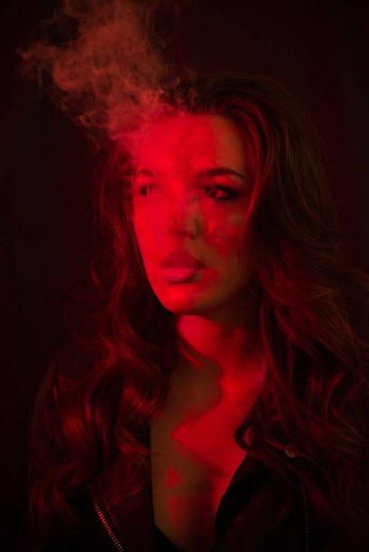Young attractive girl with long curly hair smokes an electronic cigarette and releases smoke in the red spotlights. - Foto, imagen