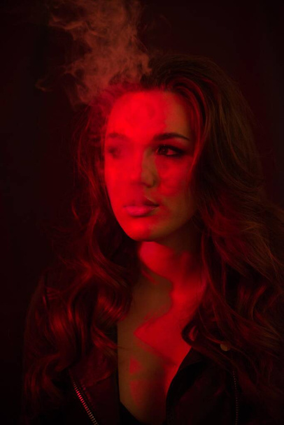 Young attractive girl with long curly hair smokes an electronic cigarette and releases smoke in the red spotlights. - Photo, image