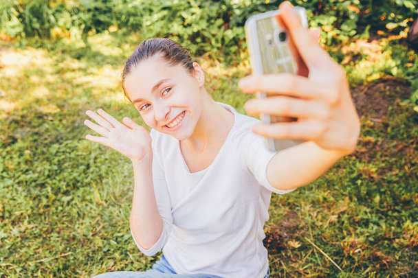 Young funny girl take selfie from hands with phone sitting on green grass park or garden background. Portrait of young attractive woman making selfie photo on smartphone in summer day - Foto, Imagem