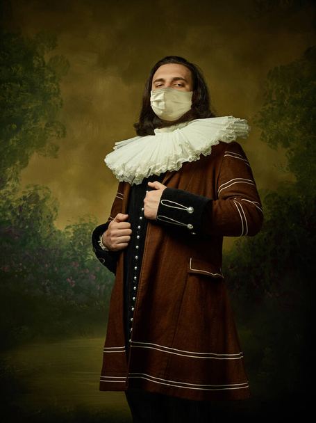 Mladý muž jako středověký rytíř na tmavém pozadí nosí ochrannou masku proti koronaviru - Fotografie, Obrázek