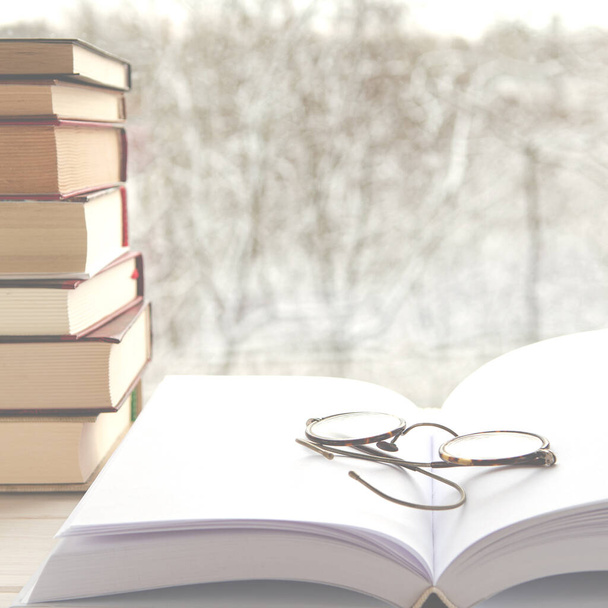 Παλιά πολύχρωμα βιβλία στέκονται σε ένα ξύλινο ράφι με φόντο το χειμερινό δάσος δίπλα σε γυαλιά ανάγνωσης - Φωτογραφία, εικόνα