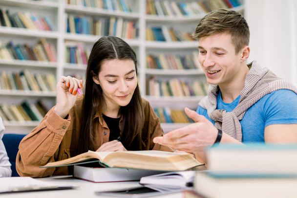 Studenten, Kaukasische knappe jongen en mooi meisje, zitten aan een tafel in een bibliotheek terwijl ze boeken lezen en samen leren, bespreken hun thuiswerk of examenvoorbereiding. Onderwijs, vriendschap - Foto, afbeelding