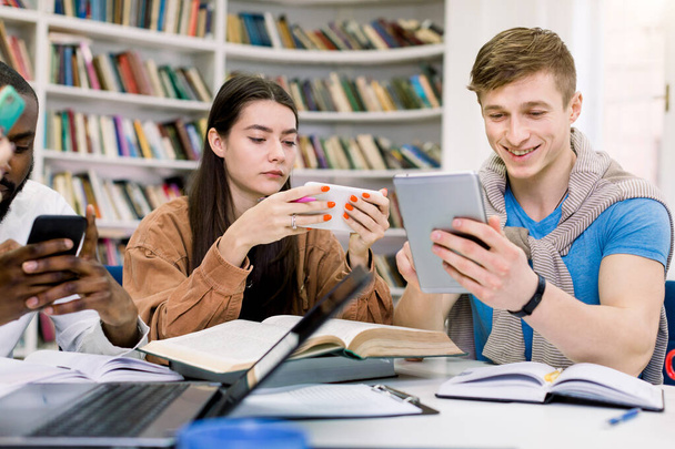 Multietničtí přátelé, vysokoškoláci studují v knihovně a používají své přístroje, smartphone a tablet, zatímco mají přestávku při přípravě na zkoušky. Knihy, notebooky a bloky na stole - Fotografie, Obrázek