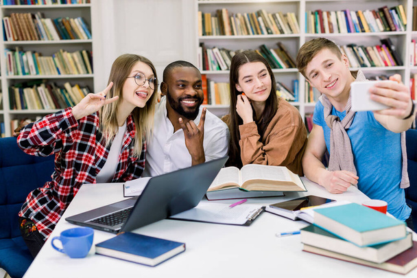 Happy young university students making selfie photo togethr while studying in library. Group of multiracial people in college library sitting together at table with books and laptop - Foto, imagen