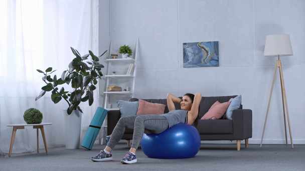 Deportista sonriente haciendo abdominales en la pelota de fitness en la sala de estar
 - Metraje, vídeo