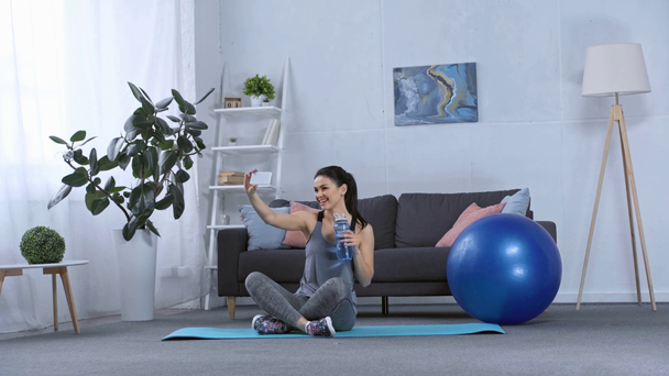 Sportswoman sorrindo levando selfie com smartphone no tapete de fitness em casa
 - Filmagem, Vídeo