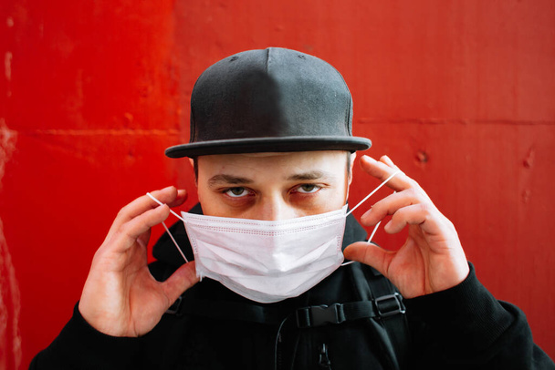 Een jongeman in zwarte sportkleding en een honkbalpet met een medisch masker op zijn gezicht bij de rode muur. bescherming tegen het virus - Foto, afbeelding