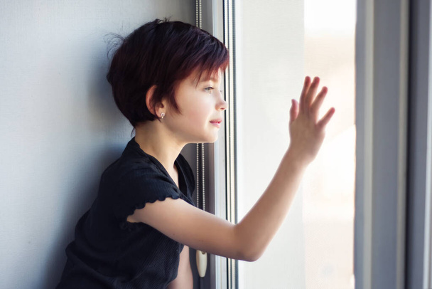 Trauriges kleines Mädchen, das aus dem Fenster schaut und Glas mit der Hand berührt. Kind während der Quarantäne wegen Coronavirus-Pandemie, das durch Fenster nach draußen starrt. Isolation und Selbstisolierung. - Foto, Bild